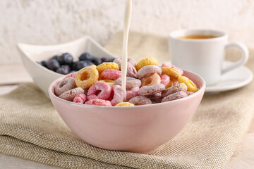 Pouring of milk into bowl with colorful cereal rings on napkin against white grunge background