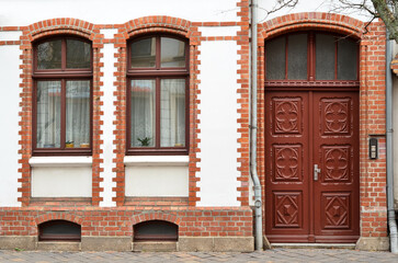 Fototapeta premium Historic building facade with arched windows and ornate wooden door, red brick details, white plaster, and paved sidewalk