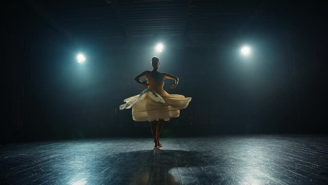 Graceful ballerina dancing elements of classical ballet on stage in backlight of spotlights. Silhouette of a young ballet dancer in pointe shoes and flowing dress