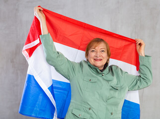 Smiling aged woman waving national tricolor flag of Netherlands posing against gray studio...