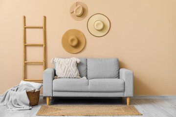 Cozy grey sofa with cushion, plaid in basket, ladder and hanging hats near beige wall