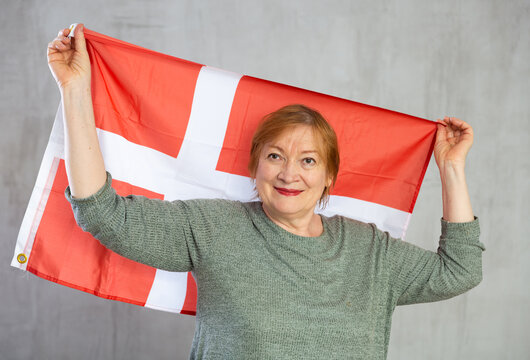 Smiling elderly woman waving national flag of Denmark while posing against gray studio background