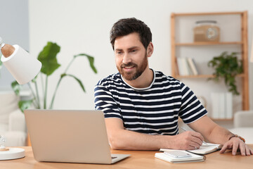Home workplace. Happy man taking notes while working with laptop at wooden desk in room