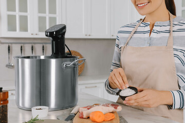 Woman salting meat near pot with sous vide cooker at table in kitchen, closeup. Thermal immersion...