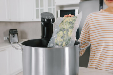 Woman putting vacuum packed broccoli into pot with sous vide cooker in kitchen, closeup. Thermal...
