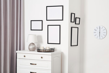 Empty frames hanging on white wall, wooden chest of drawers and lamp indoors