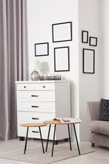 Empty frames hanging on white wall, chest of drawers and wooden tables indoors