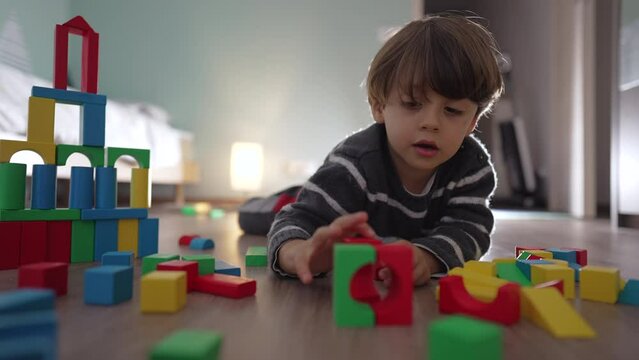 Small boy playing with toy blocks on bedroom floor. Child laying on floor plays with colorful building block construction