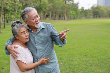 Happy asian senior man and woman walking and hugging while pointing something with picnic basket in garden outdoor. Lover couple going to picnic at the park. Happiness marriage lifestyle concept.