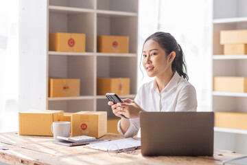 Asian SME business woman using laptop computer Phone checking customer order online shipping boxes at home. Starting Small business entrepreneur SME freelance. Online business, Work at home concept.