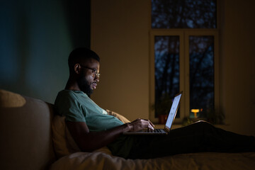 Focused African American programmer man in dark room lying on bed work on laptop. Workaholic...