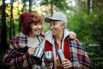 Two elderly woman friends walk in the forest, pour coffee from a thermos, have a great time together