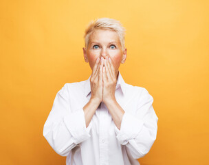 Senior grey-haired woman in white blouse covering her mouth with palms, both hands to her face