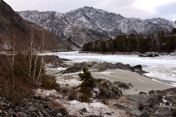 Young pine and several birch trees on the rocky bank of a beautiful frozen river flowing through a valley in the mountains with snow-capped peaks.