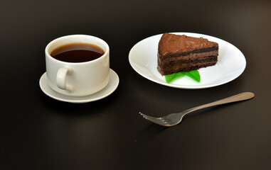 Dessert fork, a cup of hot black coffee on a saucer and a plate with a piece of chocolate layer cake with mint leaves on a black background.