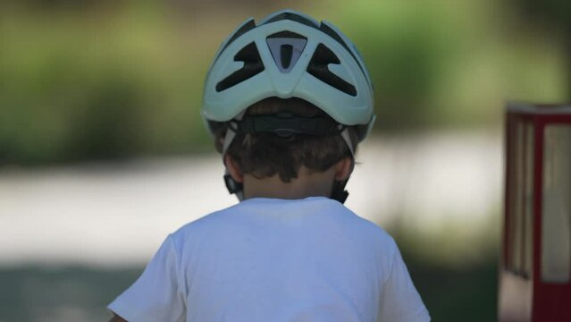 Back of cyclist child riding bike outdoors with helmet. Small boy rides bicycle at park in sunny day