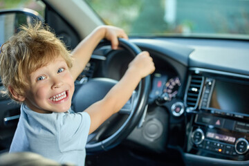 Happy boy is holding onto the steering wheel of the car, smiling broadly with joy. Smiling child in a modern car sits in the driver's seat, dreaming of winning a real race