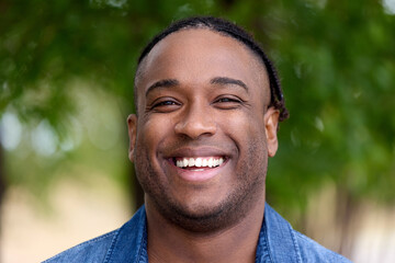 Portrait of a happy fashionable black man traveling alone through the resort town, against the backdrop of greenery in the park. Close-up of the face of a laughing tourist walking through the woods