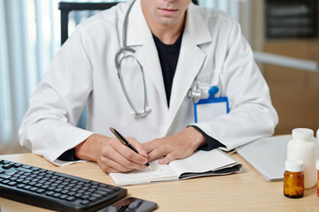 Close-up of young male doctor in lab coat making notes in medical card of online patient or writing down prescriptions during consultation