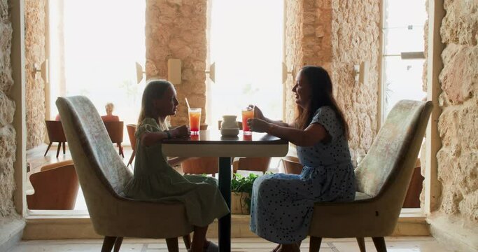 Mom and teen daughter silhouette are drinking cocktails in old restaurant with stone walls big windows, side view. Family snack in cafe. Woman teen girl talking and smiling. Having lunch together. 