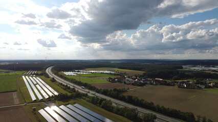 Fototapeta na wymiar Ansbach, Germany / Bavaria - August 18, 2020: Cloud and sun reflective blue solar panel energy power farm along a truck and car highway Autobahn with wind turbines in the distance view from above.