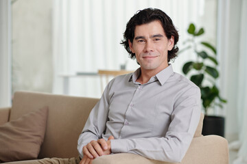 Young smiling businessman, blogger or psychologist in light blue shirt sitting on couch in home environment and looking at camera