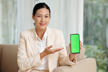 Happy young brunette businesswoman presenting new mobile application while holding smartphone with green screen and looking at camera