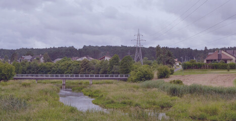 Fototapeta na wymiar Panoramic view of the swampy river, the bridge, the power line and the houses on the shore. Cloudy summer day on the river Voke in the suburbs of Vilnius, Lithuania.