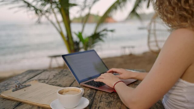 Female developer working on laptop by the ocean. Young woman freelancer coding at outdoor tropical cafe. Caucasian girl working remotely typing on computer at exotic location. Worldwide work concept.