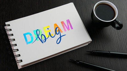 DREAM BIG colorful hand lettering in notebook with cup of coffee and pens on black wooden desk