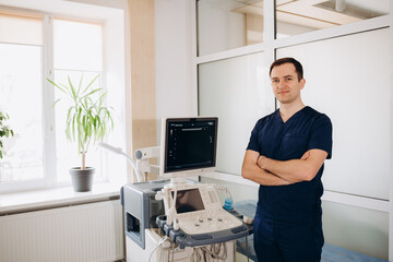 Charming young male doctor smiling to the camera, sitting at his office near ultrasound scanning machine. Handsome friendly doctor enjoying working at the hospital, copy space. Ultrasonic concept