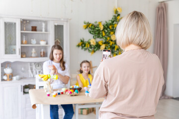 Obraz premium Mom takes pictures of two daughters sitting at kitchen at home during Easter holidays. Easter preparation coloring eggs together taking selfie photo on smartphone smiling cheerful