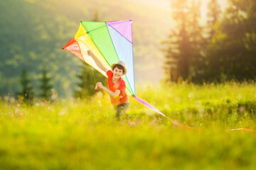 Little child boy playing multicolored rainbow kite in tall grass on a sunny summer or spring day at sunset. Active time outdoors. Mountain landscape with coniferous trees.