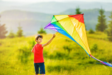 School-age boy run huge colored rainbow kite with long tail at sunset or sunrise in summertime. Mountain landscape with coniferous trees on horizon.