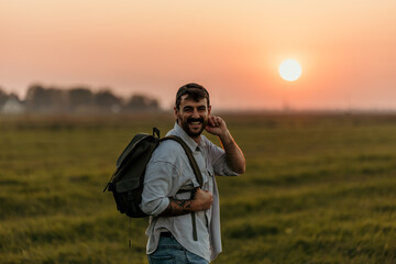 Fototapeta premium Portrait of a cheerful friendly male walking on the field with a backpack during a beautiful sunset outdoors.