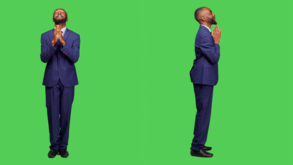 Startup entrepreneur praying to god for good luck in studio, wearing formal suit over green screen backdrop. Young male employee holding hands in a prayer sign, religious person.