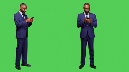 Corporate employee texting messages on mobile phone, standing over full body greenscreen in business suit. Startup manager working on smartphone and browsing online website in studio.