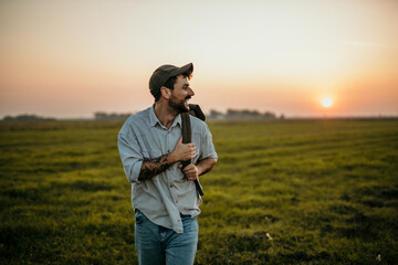 A handsome man in casual wear walks on a grassy field, having a backpack, and enjoys the fresh air. Digital nomad concept