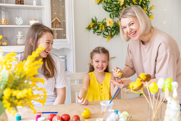 A happy mom helps her little daughter to clear paint from a hand. Preparation for the Easter holiday Small girl stained with paint while decorating easter eggs and crying at home in the kitchen.