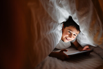 Happy boy lying under blanket and watching cartoon on tablet at night at home.