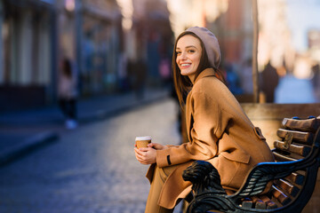 Young smiling woman girl drinking morning coffee resting on bench. Old town street background. Warm...