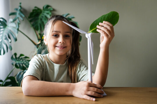 Smiling girl holding wind turbine model and leaf at home