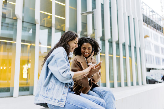 Happy young woman with friend using smart phone in front of building