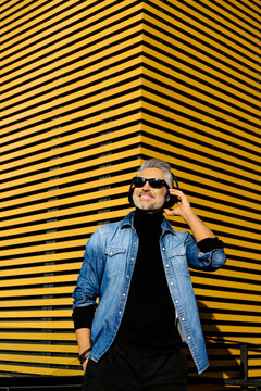 Smiling businessman wearing wireless headphones standing in front of yellow striped building