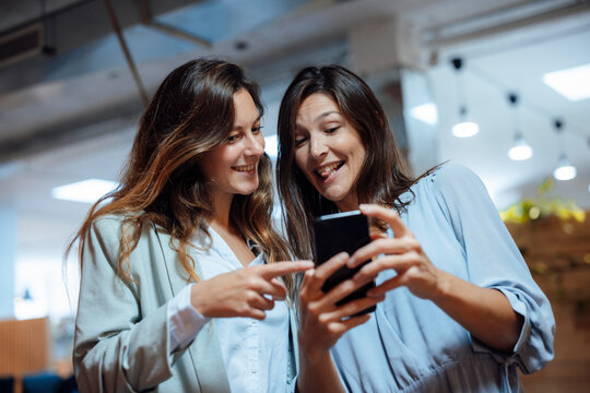 Happy businesswoman sharing mobile phone with colleague in office