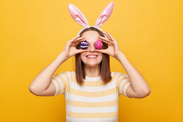 Portrait of cheerful funny woman wearing bunny ears covering eyes with easter eggs, having fun, smiling to camera, isolated over yellow background, hiding face.