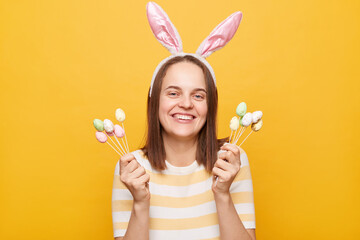 Indoor shot of charming positive attractive woman wearing bunny ears holding cake pops easter eggs isolated over yellow background, smiling to camera.