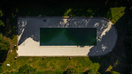 Aerial view on a swimming pool in a garden. Around the poolside there are many trees. The pool is empty and nobody is swimming.
