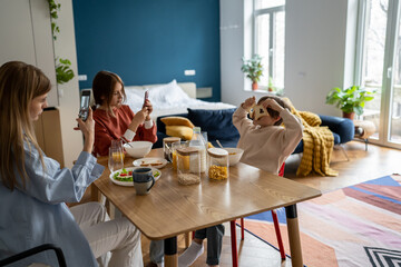 Playful boy covering eyes with bread toasts posing for photo, having fun while eating breakfast together with family members. People using smartphones during mealtime. Mobile devices and family time