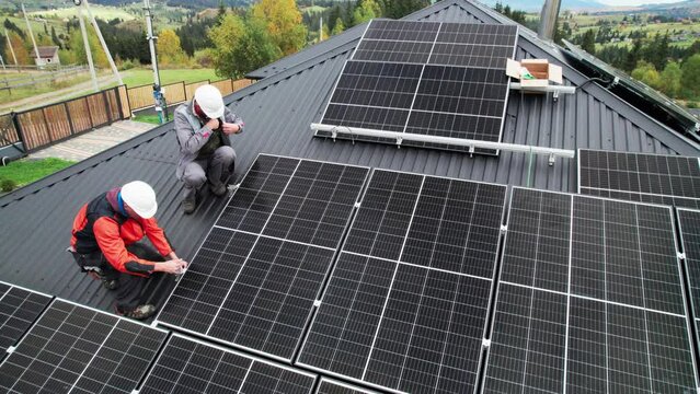 Technicians installing photovoltaic solar panels on roof of house. Drone view of men engineers in helmets building solar module system with help of hex key. Concept of alternative, renewable energy.